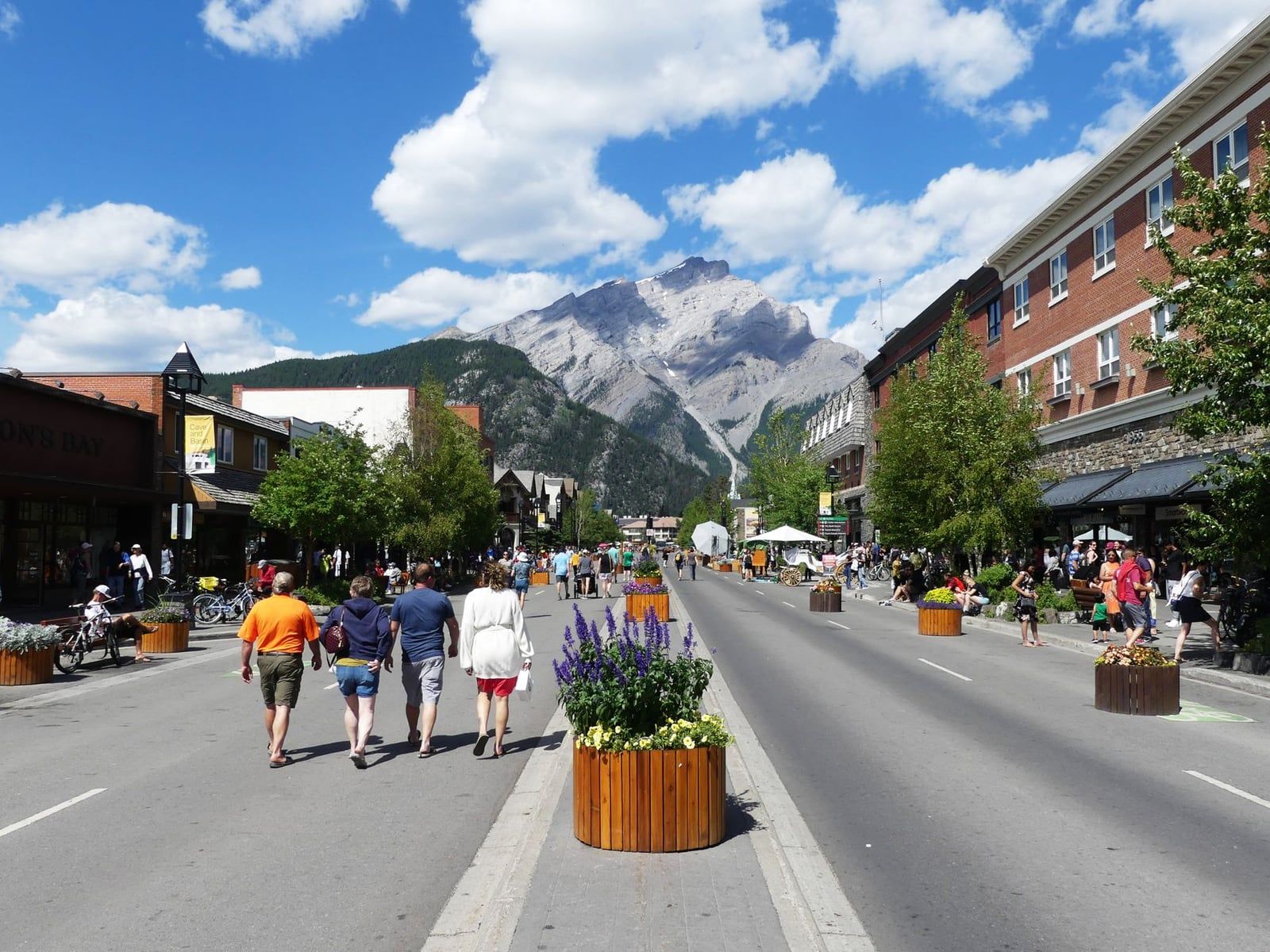 Pedestrians stroll along Banff Avenue lined with shops and flower planters, with Cascade Mountain rising in the background in Banff National Park.