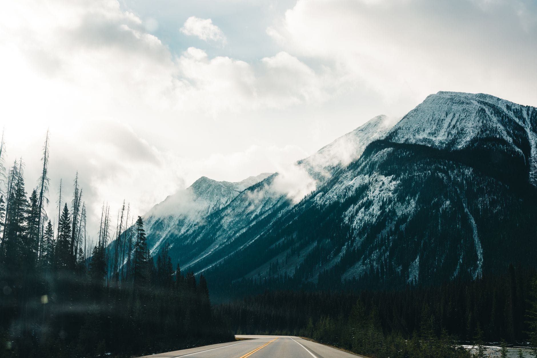 Paved highway passing through evergreen forest toward towering, snow-dusted mountains under dramatic clouds in Banff National Park.
