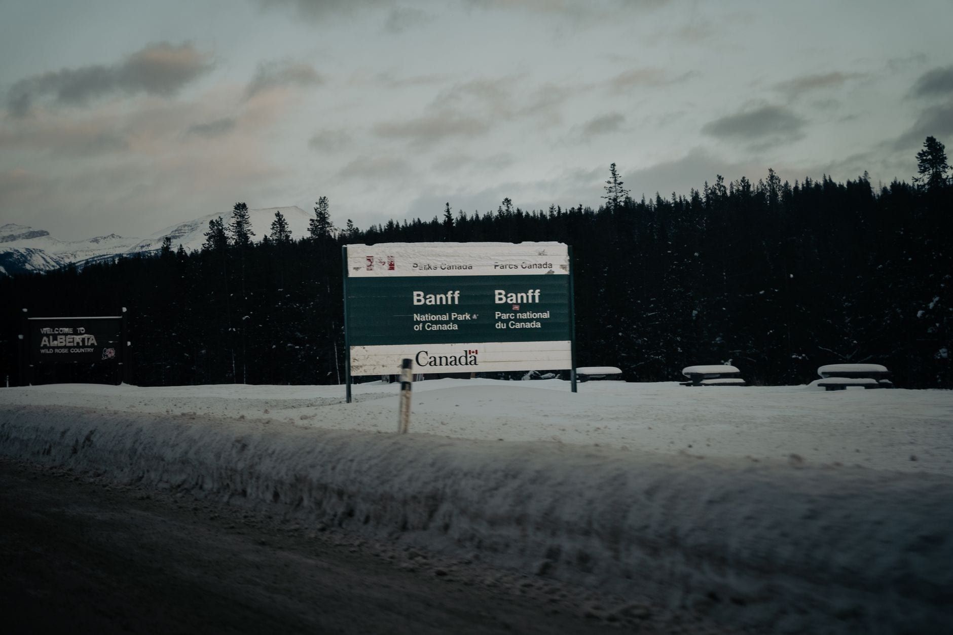 A snow-covered Parks Canada entrance sign welcomes visitors along the roadside to Banff National Park in winter.
