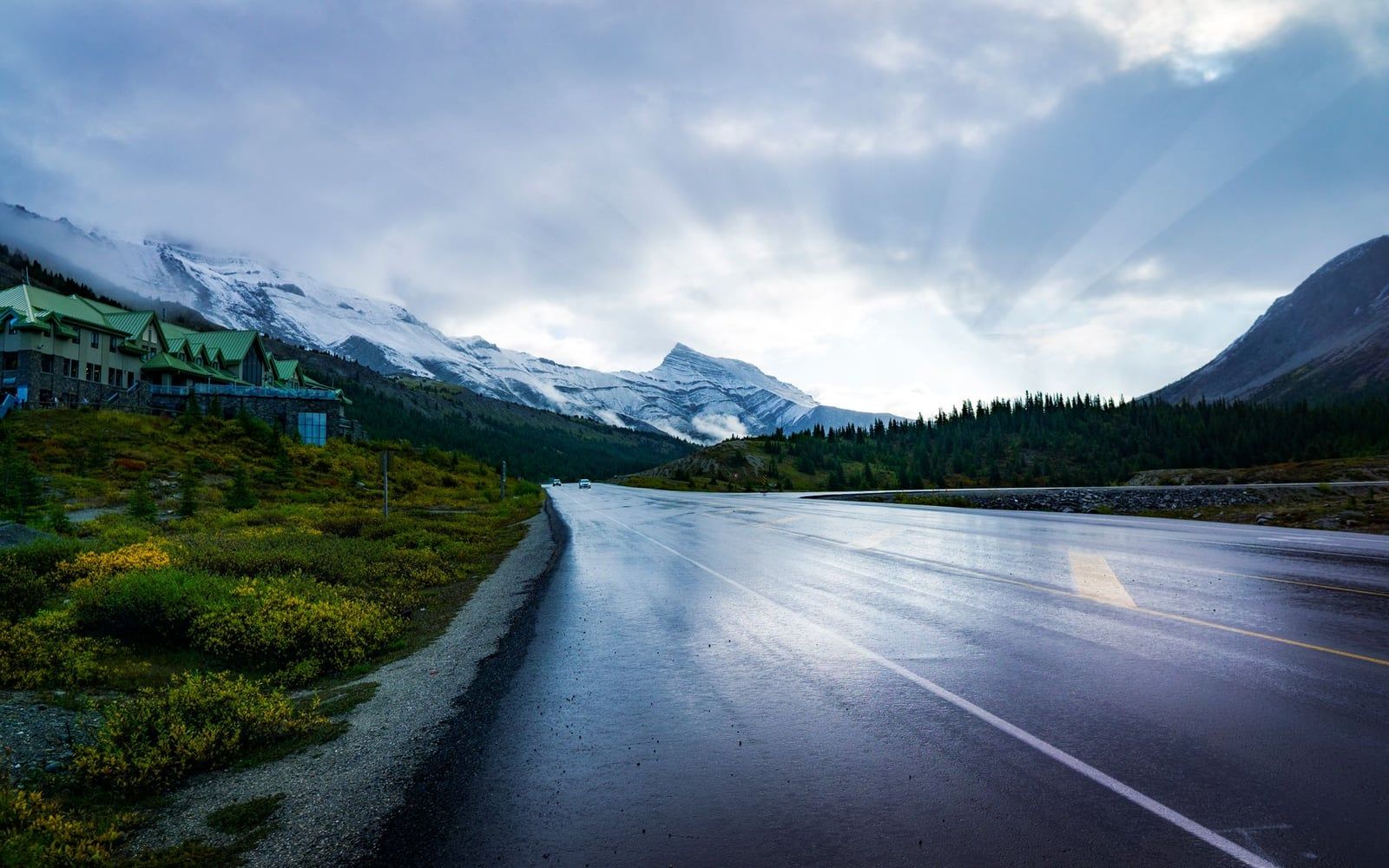 The Icefields Parkway stretches past the Columbia Icefield Discovery Centre toward snow-dusted mountains beneath a sunlit sky.