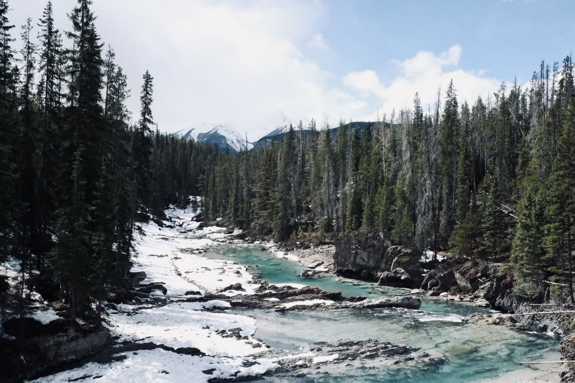 A turquoise river winds through a snowy evergreen forest toward distant mountain peaks in Banff National Park.