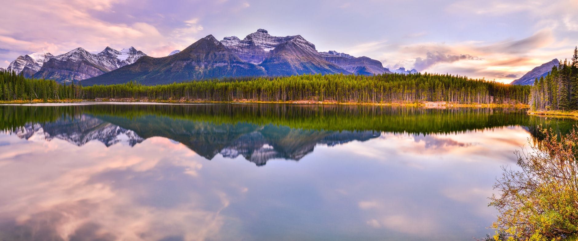 Snow-dusted mountain peaks and evergreen forests reflect across the calm water of Herbert Lake at sunset in Banff National Park.